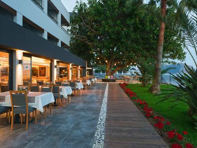 Restaurantterrasse mit gedeckten Tischen, beleuchtet, neben Holzweg und grünem Garten mit roten Blumen.