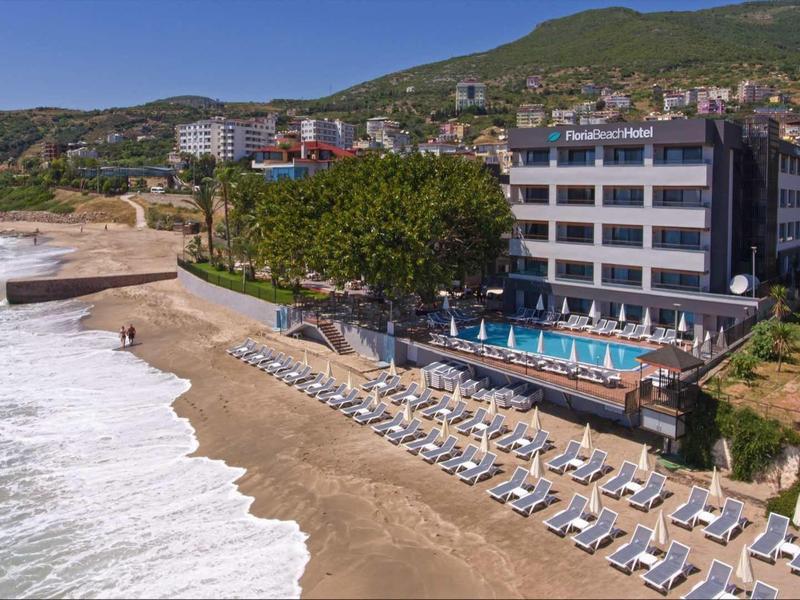 Strand mit Wellen, Liegestühlen und großem Hotelgebäude am Fuß bewaldeter Hügel.