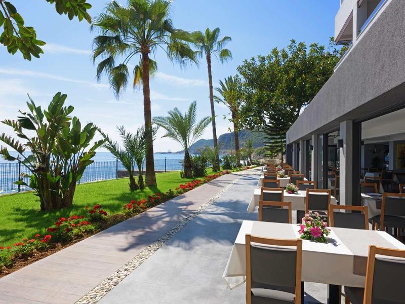 Restaurantterrasse mit gedeckten Tischen, Palmen, grünen Pflanzen und Meerblick bei sonnigem Wetter.