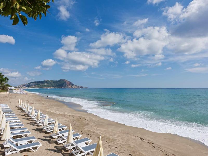 Breiter Sandstrand mit weißen Liegestühlen, blauem Meer und bewölktem Himmel im Hintergrund.