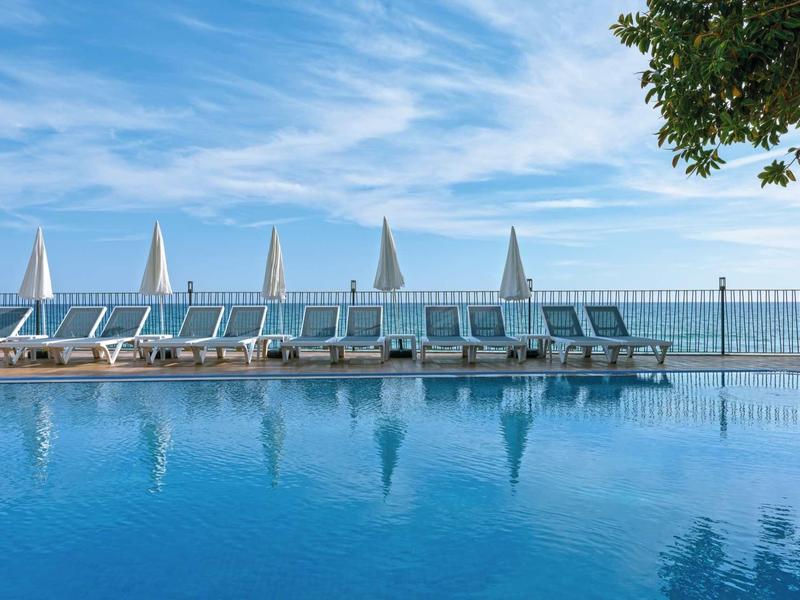 Ein ruhiger Pool mit weißen Liegestühlen und blauen Sonnenschirmen unter wolkigem Himmel.