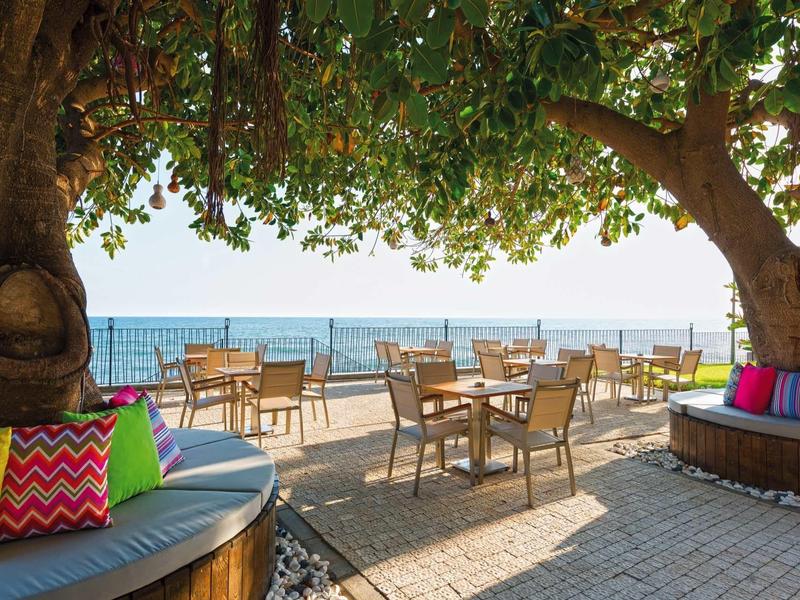 Sitzbereich mit bunten Kissen unter großen Bäumen, Blick auf Terrasse mit Tischen und Meer im Hintergrund.