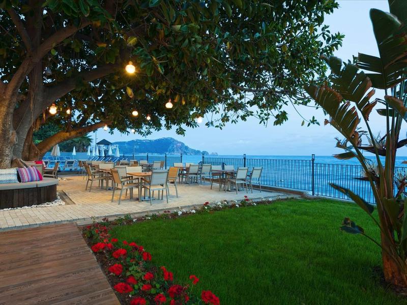 Garten mit Rasen, roten Blumen, Bäumen, Sitzbereich und Meerblick bei Abendlicht.