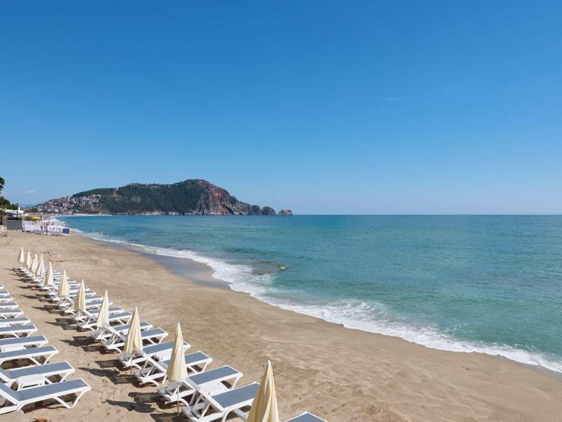 Breiter Sandstrand mit weißen Liegen, blauem Himmel und ruhigem Meer, Hügel im Hintergrund.