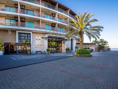Vista frontal de un hotel con balcones, palmeras y entrada pavimentada bajo cielo despejado.
