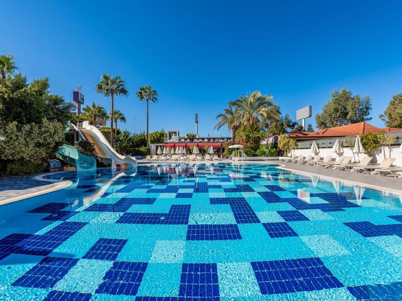 Large hotel pool with blue mosaic tiles, waterslide, and sun loungers under clear sky.