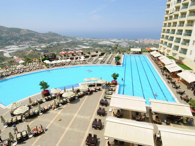 Gran piscina de hotel con tumbonas y vista a colinas y mar en el horizonte.