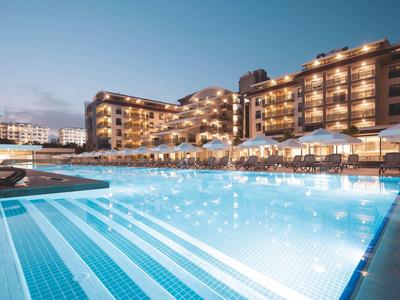 Großer beleuchteter Hotelpool bei Abenddämmerung mit Liegestühlen und mehrstöckigem Hotelgebäude.