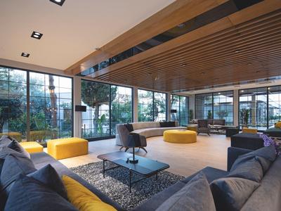 Modernes Wohnzimmer mit großen Fenstern, grauen Sofas, gelben Sitzgelegenheiten und Holzbalkendecke.