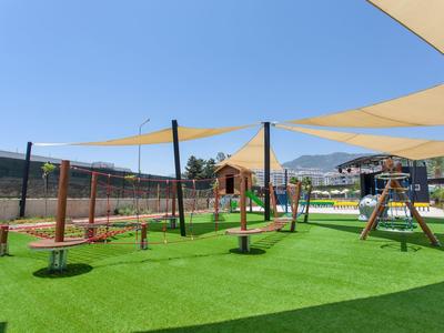 Spielplatz mit grünem Kunstrasen, Klettergerüst, Schaukeln und Sonnensegeln unter blauem Himmel.