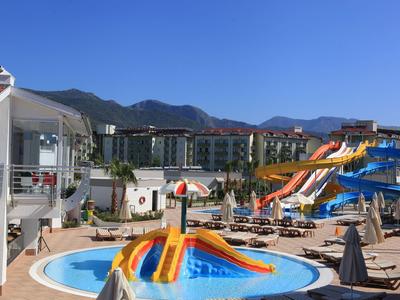 Wasserpark mit bunten Wasserrutschen, Pools, Liegestühlen und Bergen im Hintergrund unter blauem Himmel.