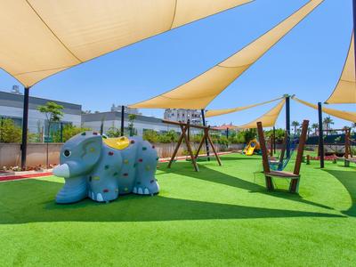Kinderspielplatz mit grünem Kunstrasen, blauem Dinosaurier-Rutsche und Holzschaukel unter Sonnensegeln.