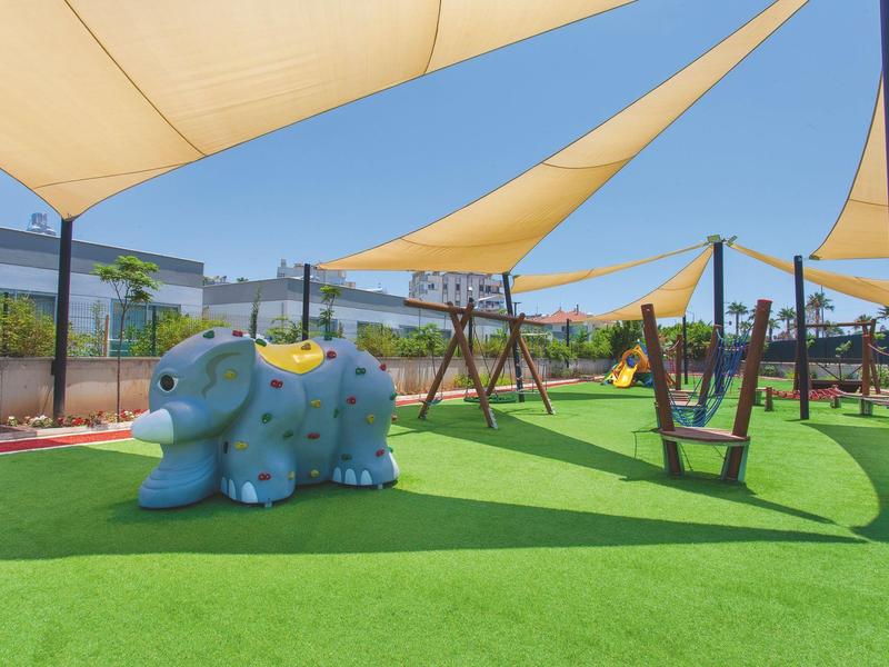 Une aire de jeux ensoleillée avec gazon artificiel vert, balançoires et un cadre d'escalade dinosaure bleu.