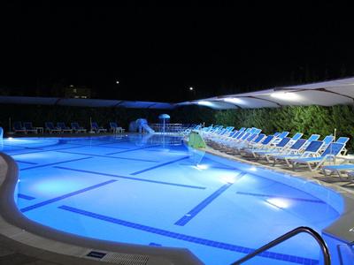 Großes beleuchtetes Schwimmbecken mit Liegestühlen und Sonnenschirmen in der Nacht.