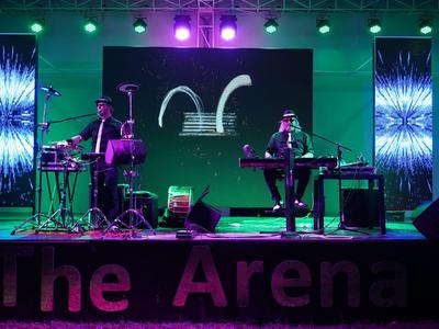 Twee muzikanten op het podium met instrumenten voor een verlichte achtergrond in The Arena.
