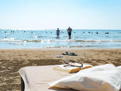 Liege mit Kissen am Sandstrand, zwei Personen im Meer und Schwimmer im Hintergrund