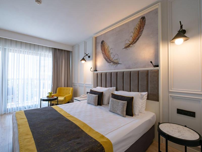 Modernes Hotelzimmer mit großem Bett, gelben und grauen Akzenten und großen Fenstern.