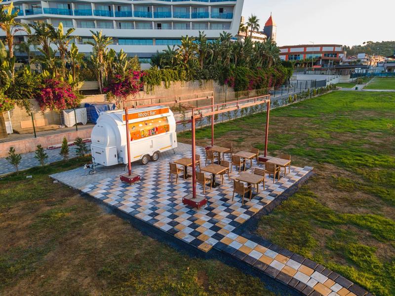 Freifläche mit Schachbrettboden, Tischen, einem kleinen weißen Anhänger und Palmen im Hintergrund.