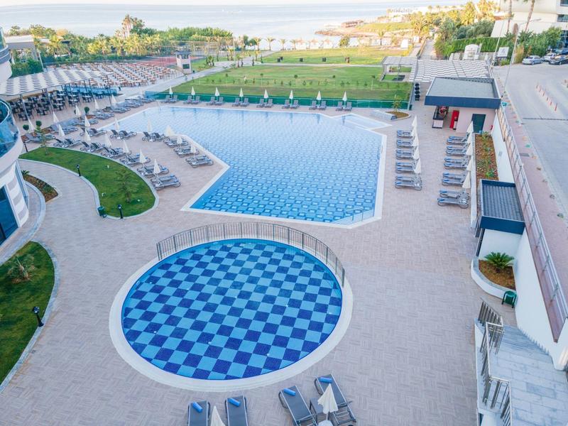 Blick auf ein modernes Hotel mit rechteckigem und rundem Pool, umgeben von Liegestühlen und Grünflächen.