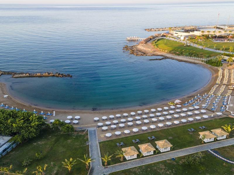 Halbmondförmiger Sandstrand mit vielen Sonnenschirmen, Palmen und angrenzender grüner Parkanlage.