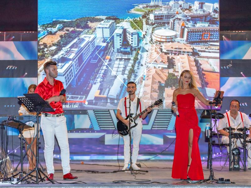 Band mit vier Musikern spielt vor großem Bildschirm mit Stadt- und Meeresansicht, helle Bühne.