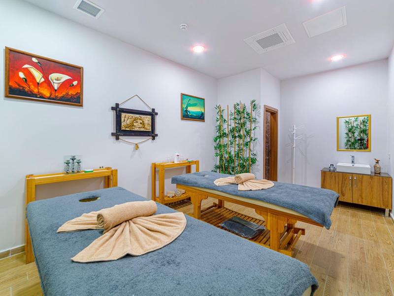 Ein heller Raum mit zwei blauen Massageliegen, Holzmöbeln und Pflanzen an weißen Wänden.