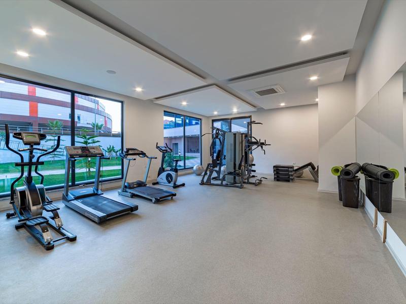 Heller Fitnessraum mit Laufbändern, Crosstrainern, Kraftgerät und großen Fenstern zum Außenbereich.