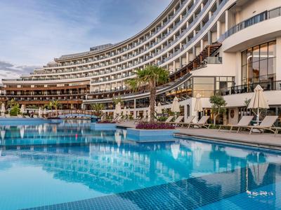 Nowoczesny kompleks hotelowy z dużym basenem, leżakami i palmami pod pochmurnym niebem.