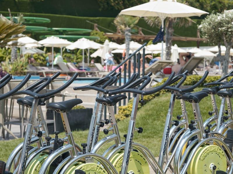 Several stationary bikes outdoors on a sunny day near umbrellas and pool area.