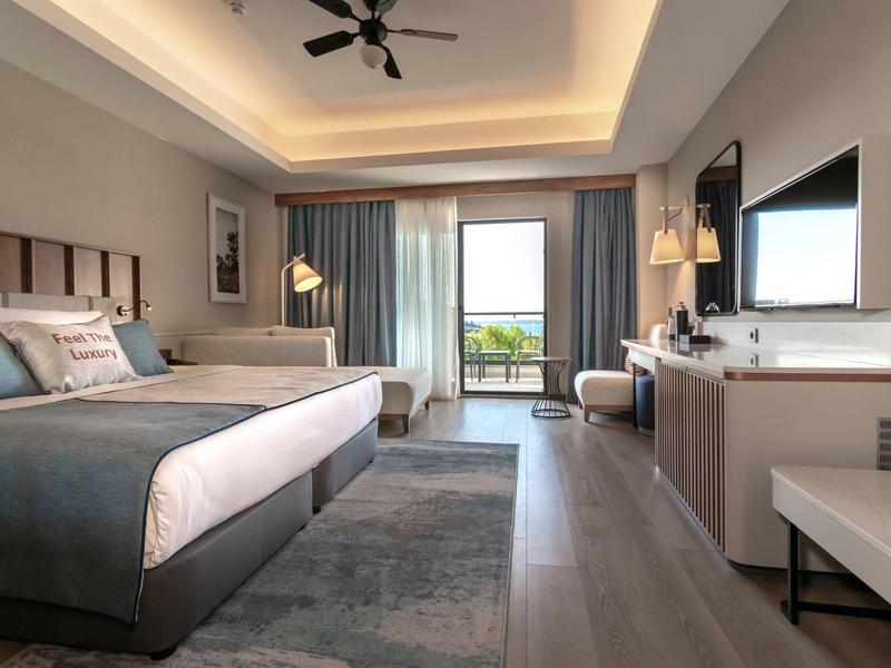 Modern hotel room with large bed, TV, desk, and balcony view.