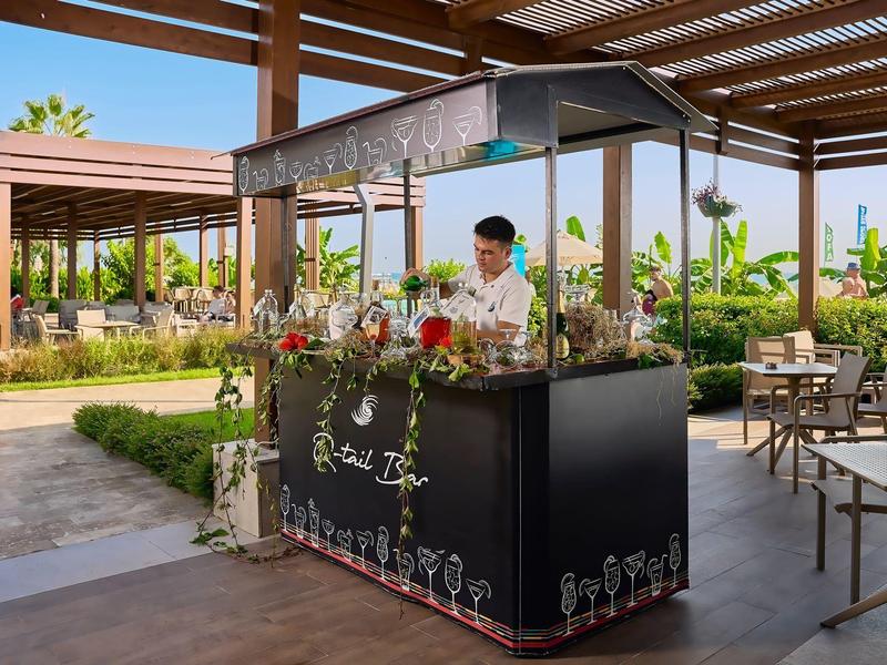 Bar with a bartender under a covered outdoor hotel area, surrounded by plants.