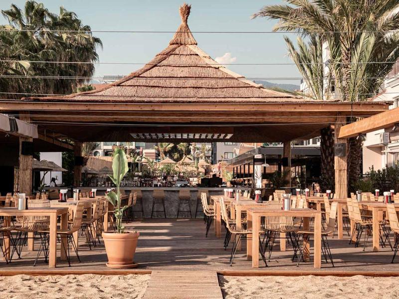 Strandrestaurant onder een groot houten paviljoen met tafels en stoelen.