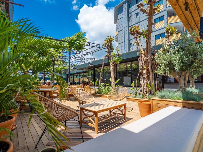Ruim terras met houten meubels, planten en moderne hotelarchitectuur onder een heldere blauwe lucht.