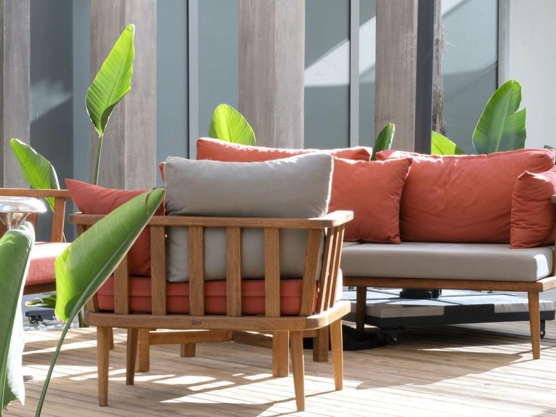 Moderne Terrasse mit Holzmöbeln, grauen Polstern, roten Kissen und grünen Pflanzen im Sonnenlicht.