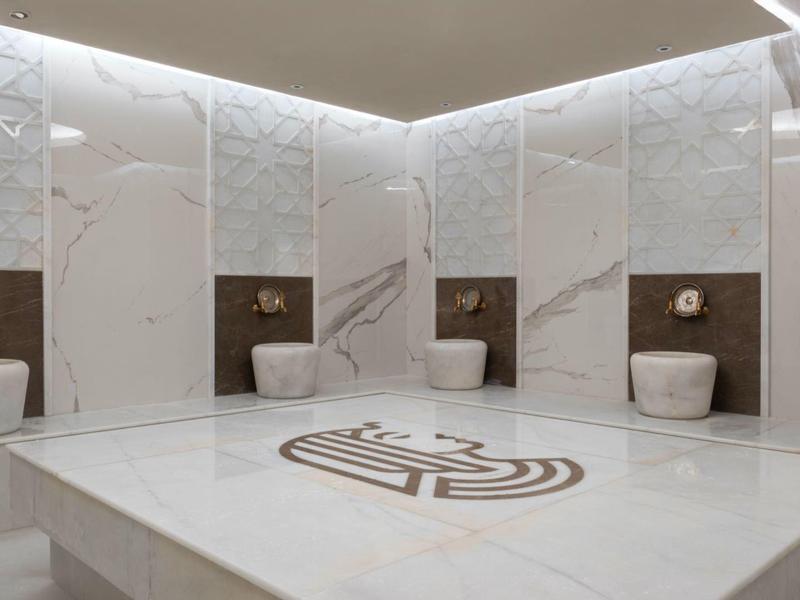 Moderner weißer Hammam mit Marmorfliesen, großen Sitzflächen und eingelassenen Wasserhähnen.