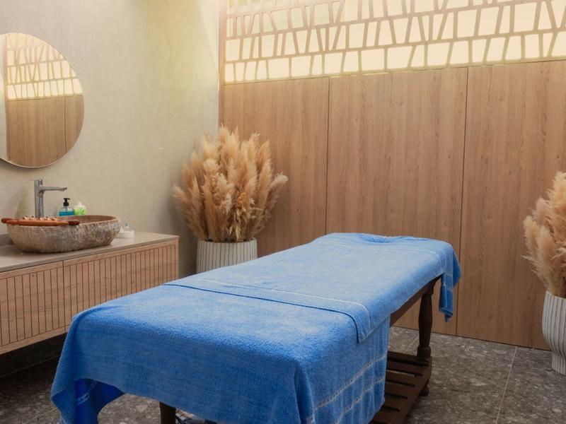 Behandlungsraum mit blauer Liege, Holzvertäfelung, Spiegel, Waschbecken und Deko mit Trockenpflanzen.