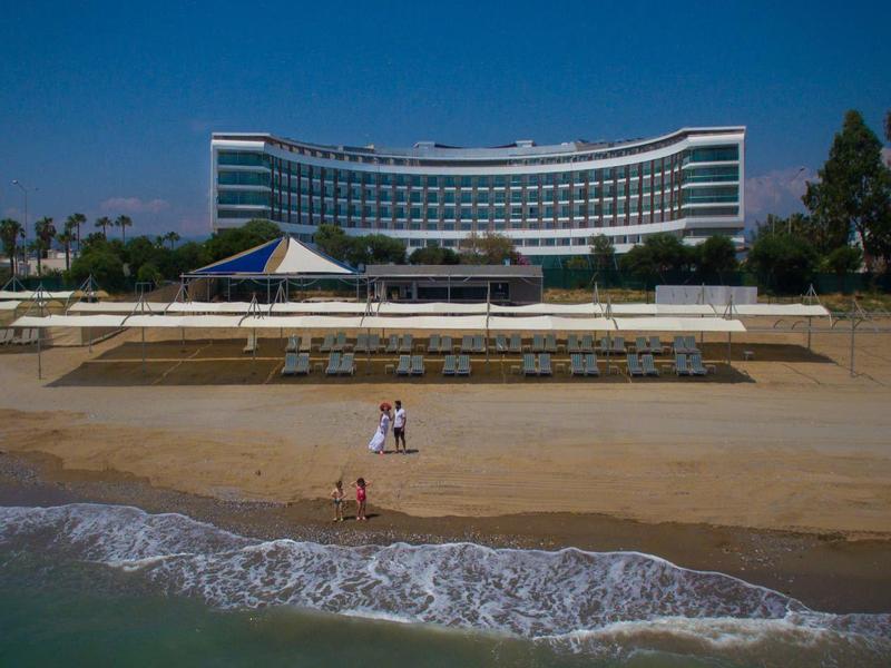 Edificio de hotel moderno en la playa con sombrillas y palmeras al fondo bajo cielo azul.