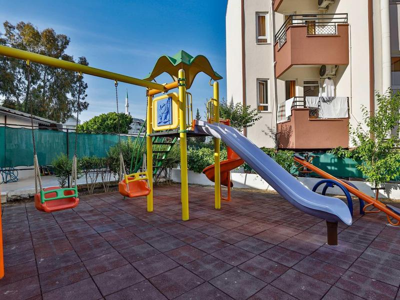 Aire de jeux pour enfants avec balançoires colorées et toboggan bleu à côté d'un immeuble résidentiel.