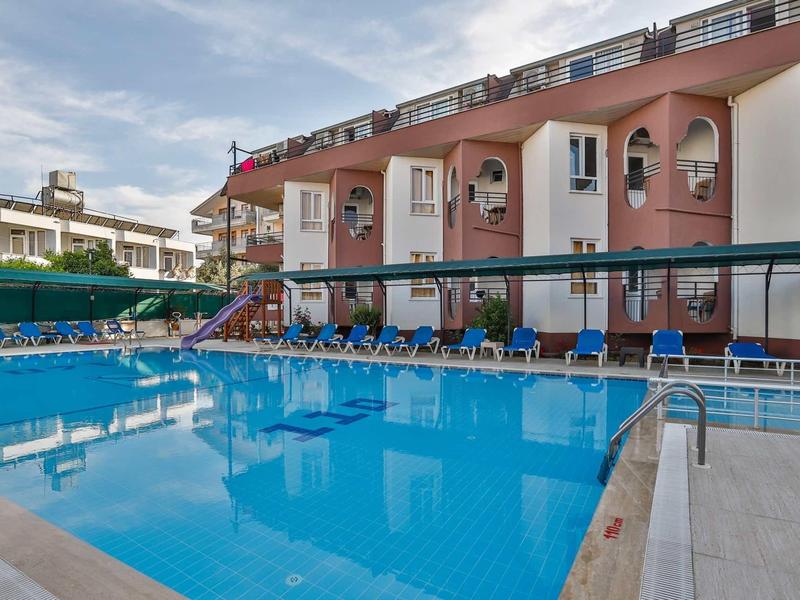 Grande piscine extérieure devant un bâtiment d'hôtel rose avec des chaises longues bleues et des parasols.