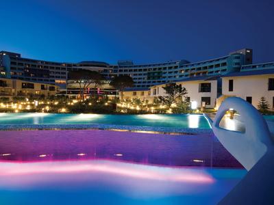 Modernes Hotel in U-Form mit beleuchtetem Pool in Blau- und Lilatönen bei Nacht.