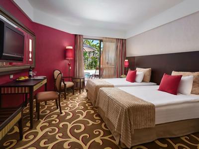 Hotelzimmer mit zwei Betten, Teppichboden, Fernseher und Balkon mit Pflanzenblick.