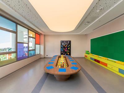 Moderner Klassenraum mit großem Holztisch, bunten Sitzplätzen, grünem Whiteboard und großen Fenstern.