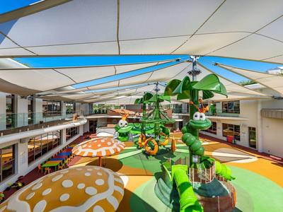 Bunte Indoor-Spielhalle mit großen Pilzen, Rutschen und grünen Klettergerüsten unter weiß-blauem Tuchdach.