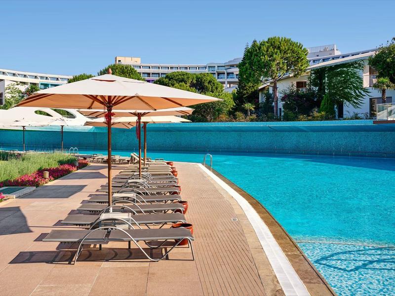 Poolbereich mit Liegestühlen und Sonnenschirmen neben klarem blauem Wasser und moderner Architektur.