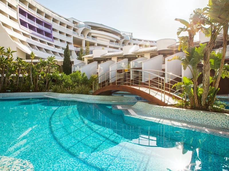 Blick auf einen Pool mit Brücke und modernen Hotelgebäuden im Hintergrund unter klarem Himmel.