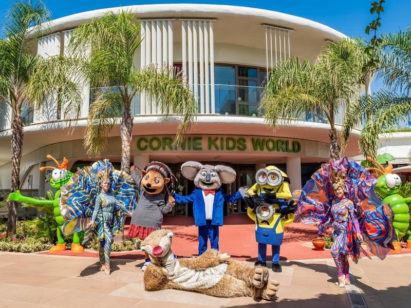 Fröhliche Kostümpersonen vor dem Eingangsbereich eines farbenfrohen Freizeitparks bei sonnigem Wetter.