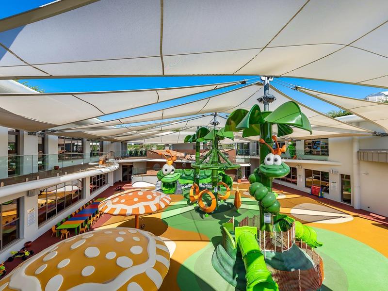 Bunter Innenhof mit Pilz-Sitzgelegenheiten und grüner Spielplatzstruktur unter einem weißen Sonnensegel.