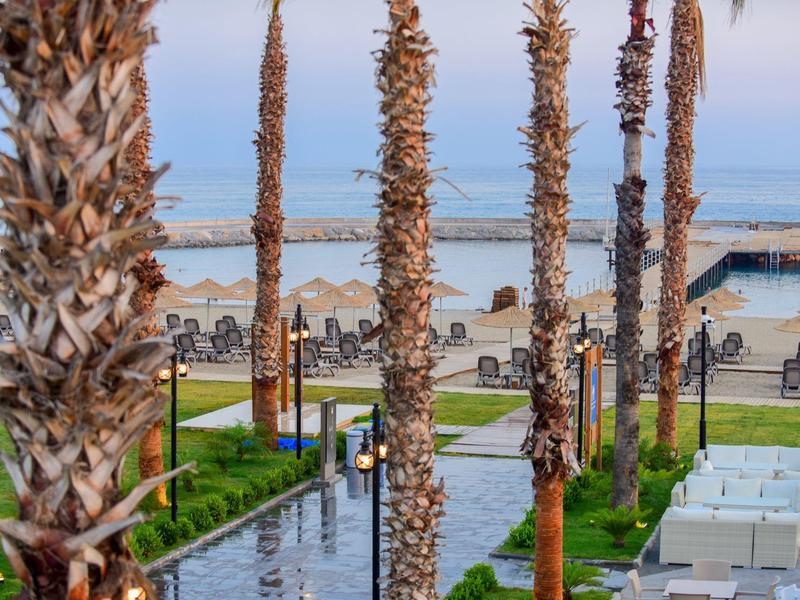 Vista de un jardín de hotel con palmeras, piscina y tumbonas junto a la playa de arena y un muelle.
