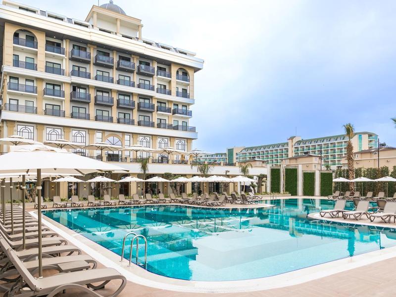 Gran piscina al aire libre con tumbonas y sombrillas frente a un edificio de hotel de varios pisos.