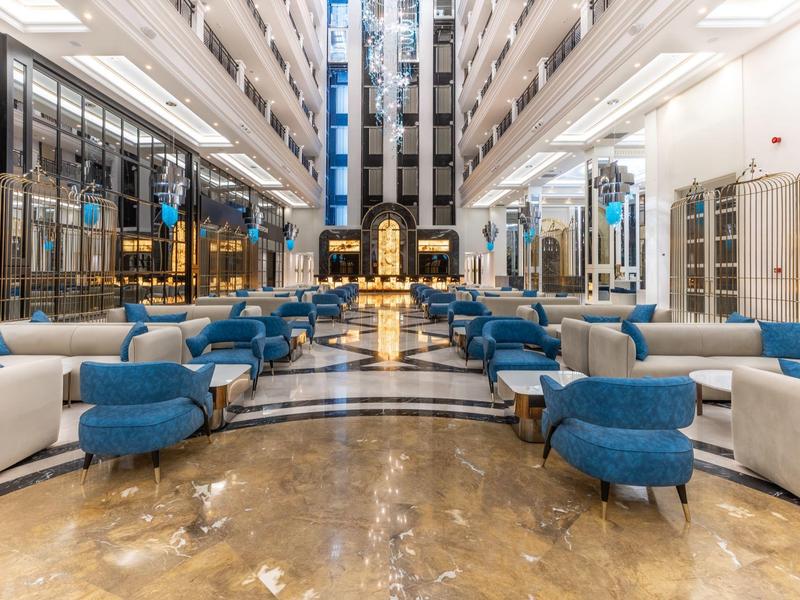 Amplio vestíbulo de hotel con sillones azules, sofás y suelo de mármol brillante bajo luz brillante.